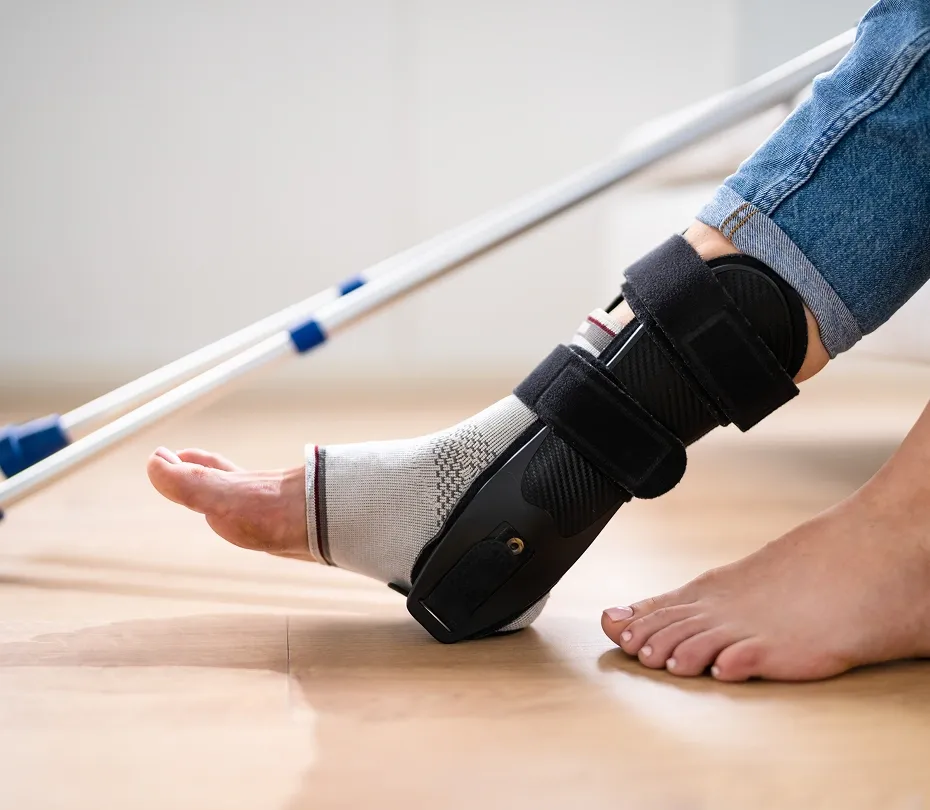Close-up of a person wearing a rigid black medical ankle brace over a sock, with crutches resting in the background.