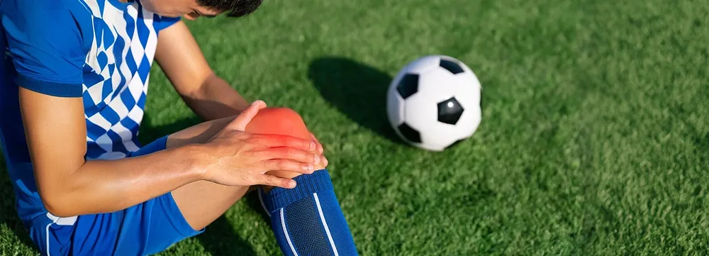 Football player sitting on the grass holding his painful knee after a sports injury with a football beside him.