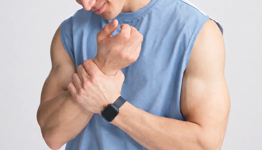 A muscular man wearing a blue sleeveless shirt and smartwatch holding his wrist in pain against a white background.