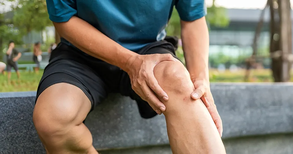 Person holding a painful knee while sitting outdoors, illustrating knee pain or a possible knee injury.