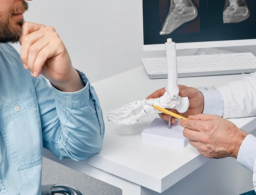 A doctor pointing a yellow pencil at an anatomical foot and ankle bone model during a medical consultation with a male patient.