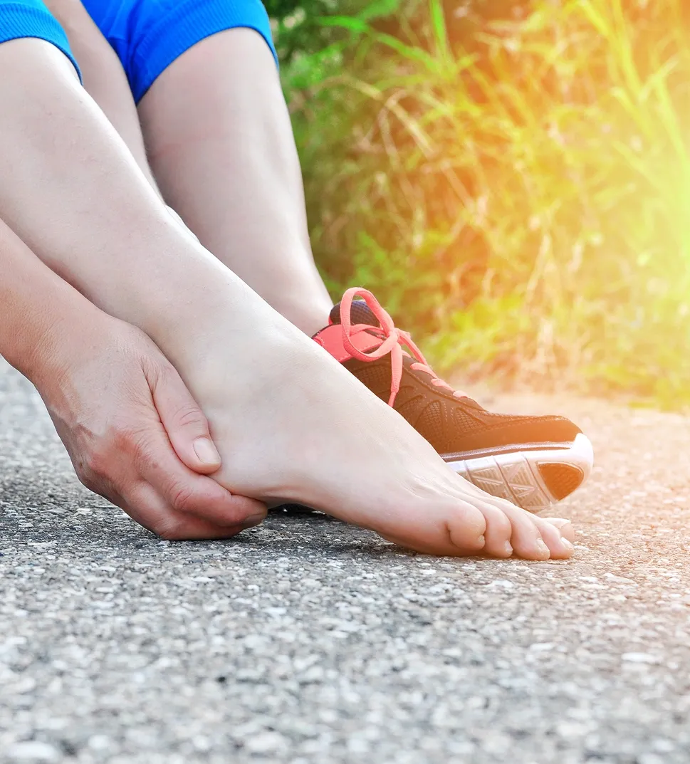 A runner sitting bare-foot on an outdoor path holding a painful heel, with a running shoe visible in the background.