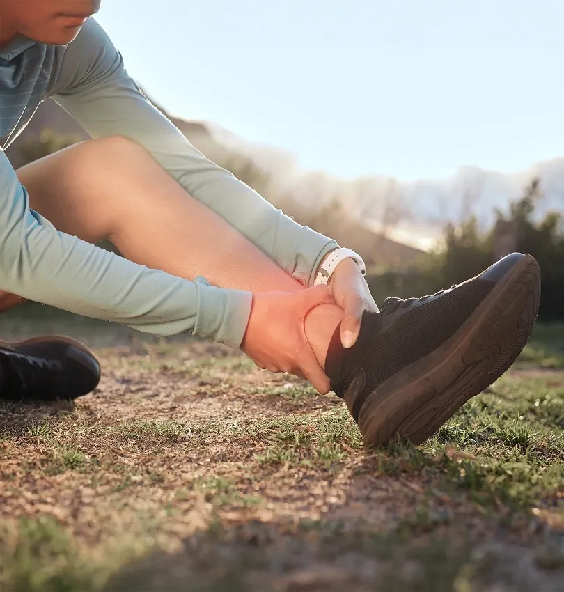 An athlete wearing a long-sleeve shirt and black running shoes sitting on the ground clutching a painful ankle injury.